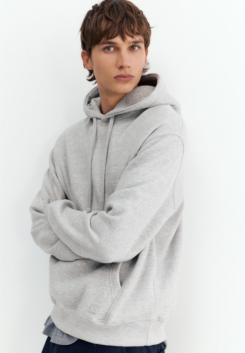 Jeune personne aux cheveux courts bruns portant un sweat à capuche gris clair, debout, les bras croisés sur un fond blanc uni.