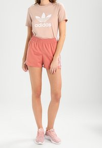 Jasnoróżowa koszulka Adidas z białym logo, połączona z koralowymi szortami z elastycznym paskiem i białymi paseczkami po bokach. Lekka tekstura.