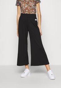 Svarta vida byxor i mjukt tyg, med hög midja och en liten logotyp på sidan. Bärs med vita sneakers.