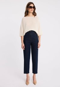 Kobieta nosząca oversize'owy kremowy sweter, ciemne jeansy z wysokim stanem, beżowe szpilki i duże okrągłe okulary przeciwsłoneczne w muszkatołowym kolorze, na białym tle.