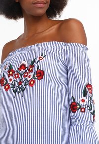 Sininen ja valkoinen raidallinen olkaimeton paita, jossa on brodeerattuja kukkakuviota punaisena, vaaleanpunaisena ja vihreänä hihoissa ja rinnassa. Pehmeä kangas.