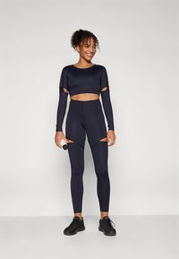 Mørkeblå atletiksæt med en cropped langærmet top med udskæringsdetaljer og højttaljede leggings med åbninger i siderne. Teksturen virker glat.