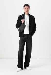 Black zip-up jacket over a white shirt, paired with dark wide-leg jeans and white sneakers. A small black crossbody bag is worn.