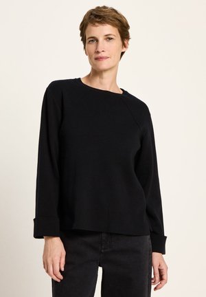 Frau mit kurzem braunem Haar, die einen schwarzen, langärmeligen Pullover und schwarze Hosen trägt, steht vor einem schlichten hellen Hintergrund.