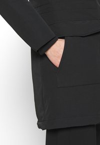 Zwarte jas met een slim fit, zachte textuur en een borstzak. De mouwen zijn voorzien van geribbelde manchetten, met een rechte zoom aan de onderkant.