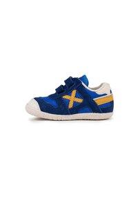 Zapatilla infantil de ante azul y marino con tiras de Velcro, suela de goma blanca y acentos amarillos, que presenta un patrón en cruz en el lateral.