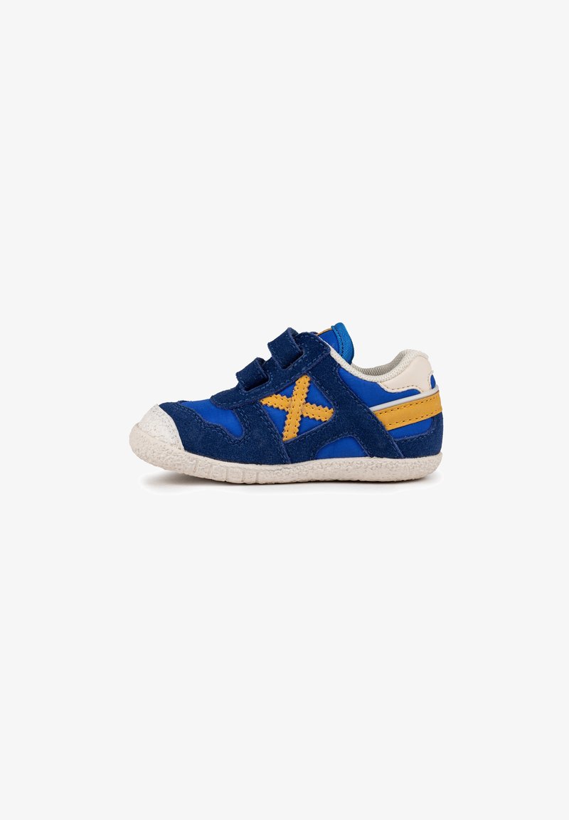 Zapatilla infantil de ante azul y marino con tiras de Velcro, suela de goma blanca y acentos amarillos, que presenta un patrón en cruz en el lateral.