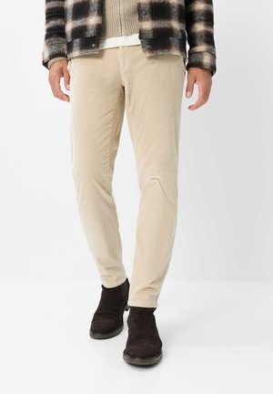 Uomo che indossa pantaloni di velluto beige, una giacca marrone a quadri e stivali scamosciati marrone scuro, in piedi su uno sfondo bianco.