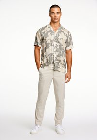 Camisa de manga corta con estampado floral en tonos verde apagado y beige, combinada con pantalones color crema y zapatillas blancas. Diseño de botones sencillo.