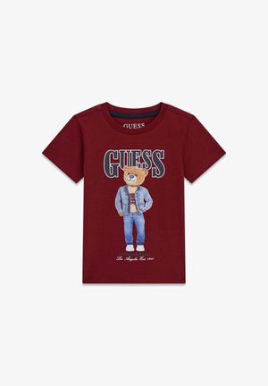 Tricou de bumbac maro roșcat cu un grafic cu urs purtând o jachetă de denim și blugi. Textul spune „GUESS” și „Authentic Denim Brand.”