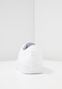 Vit canvas-sneaker med slät textur, rund tå, dragflik baktill och gummisula. Minimalistisk design, utan synliga mönster eller accenter.