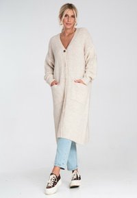 Blondynka w długim beżowym kardiganie z guzikami, jasnoniebieskich dżinsach i sneakersach na platformie w panterkę, stojąca z dłońmi w kieszeniach.