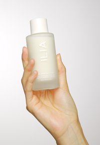 Przezroczysta szklana butelka z białą nakrętką, oznaczona napisem "ILIA THE BASE FACE MILK." Zawiera jasną ciecz; trzymana w dłoni na jasnym tle.