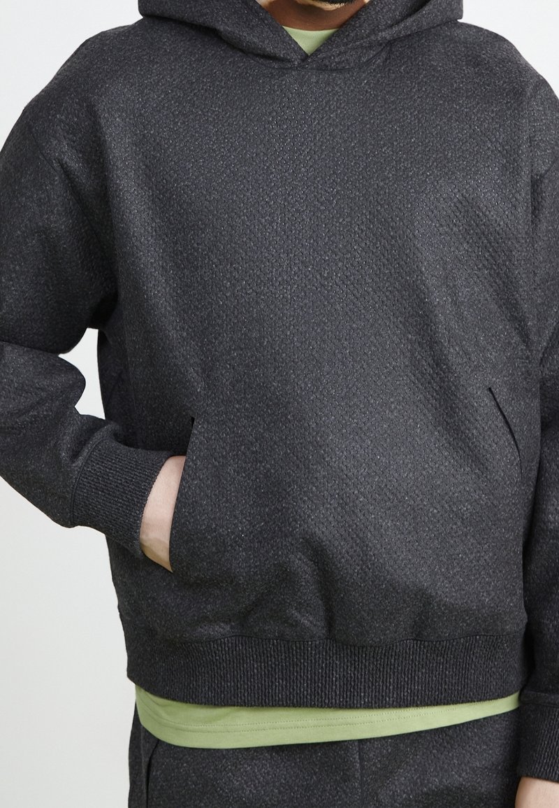 Sudadera con capucha negra hecha de tela texturizada, con un bolsillo canguro, puños y dobladillo acanalados. Lleva puesta sobre una camisa verde.