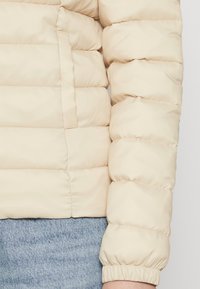 Lys beige pufferjakke med quiltet design og ribbede mansjetter. Myk, glatt tekstur; har sidelommer og elastisk hem.