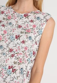 Květinová blůza na bílém pozadí, se vzory růžových, modrých a zelených květin, krátkými rukávy a malým knoflíkovým detailem na výstřihu.