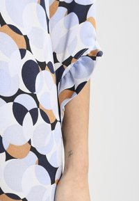 Krátký rukáv, vzorovaná blůza ve světle modré, bílé a zemitých tónech. Obsahuje kulaté tvary a strukturovanou tkaninu.