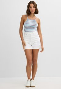 Mujer de pie con pantalones cortos blancos, camiseta sin mangas a rayas azules y zapatillas blancas frente a un fondo claro y liso.
