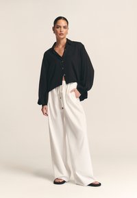 Črna bluza s srajčnim zapenjanjem in prevelikimi rokavi, združena z belimi ohlapnimi hlačami z vrvico in dekorativnimi gumbi.