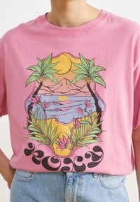 Vaaleanpunainen puuvillat-paita, jossa on trooppinen grafiikka, jossa on palmupuita, vuoria ja ranta, jota korostavat värikkäät kukkakuviot alareunassa.
