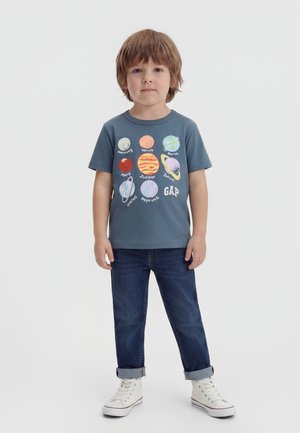 Μικρό παιδί που φορά μπλε μπλουζάκι με πολύχρωμο σχέδιο ηλιακού συστήματος, σκούρο τζιν και λευκά ψηλά αθλητικά παπούτσια σε απλό φόντο.