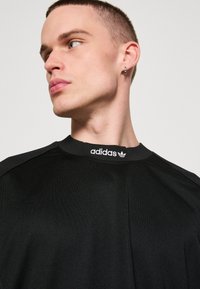 Fekete sportpóló kerek nyakkivágással és bordázott gallérral, a mellkasán egy minimalista fehér Adidas logóval. Puha, textúrált anyag.