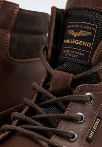 Brown leather and suede ankle boots with black textile accents, metal eyelets, and branded patch. Features textured surfaces and laces.