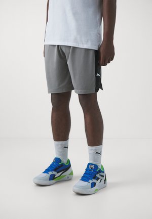 Mand iført grå Puma-shorts, hvide Puma-strømper og blå, hvide, sorte og grønne Puma-sneakers står på en hvid gulv.