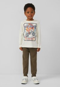 Weißes Langarmhemd mit einem Zirkus-Tiger-Graphic, kombiniert mit braunen Hosen mit seitlichen Streifen und weißen Sportschuhen.