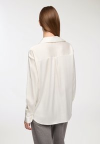 Blusa lunga a maniche con texture satinata di un beige chiaro, caratterizzata da un colletto a rever e un dettaglio con piega sul retro.