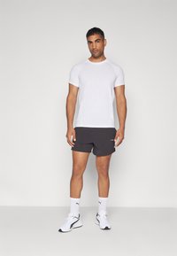 Valge lühikeste varrukatega spordipe-shirt, mis on kombineeritud tumedate lühikeste pükstega, valgete sokkide ja mustade jooksujalatsitega. Minimalistlik disain, ilma mustriteta.