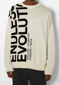 Pull en tricot beige de coupe décontractée, avec un grand texte imprimé en noir disant "ENDLESS EVOLUTION" sur le devant, et des poignets et ourlet côtelés.