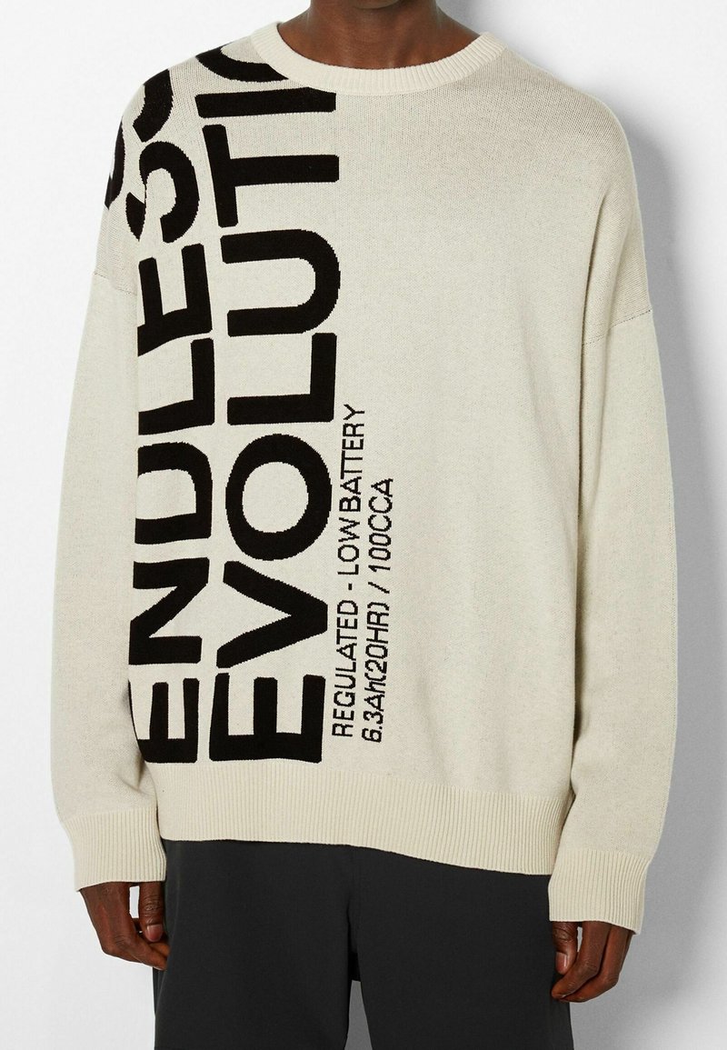 Pull en tricot beige de coupe décontractée, avec un grand texte imprimé en noir disant "ENDLESS EVOLUTION" sur le devant, et des poignets et ourlet côtelés.