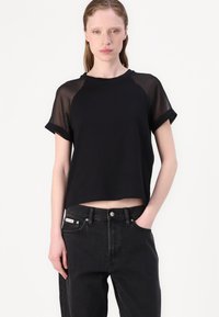 Black short-sleeve top with sheer fabric on shoulders, cropped design, and round neckline. Paired with high-waisted black jeans.