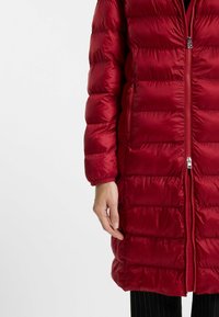 Röd puffjacka med dragkedja fram, ribbade ärmslut och vertikalt kviltade paneler. Har sidofickor och en glansig yta.