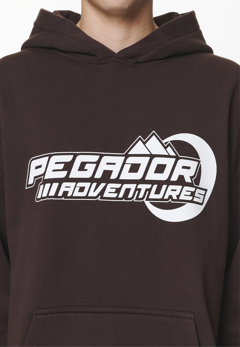 Brun hoodie mit einem weißen Grafiklogo, das Berge und den Text "PEGADOR ADVENTURES" zeigt. Hergestellt aus weichem Stoff und mit einer Fronttasche.