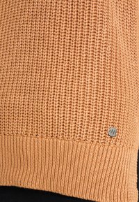 Textured, gebreide trui in licht perzikkleur met geribbelde zoom en een klein zilveren hardwaredetail met een 'N' logo aan de zijkant.