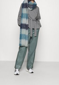 Maglione grigio a maglia con design cintato, abbinato a pantaloni in finta pelle verde acqua e una sciarpa a quadri blu, completato con scarpe da ginnastica in bianco e nero.