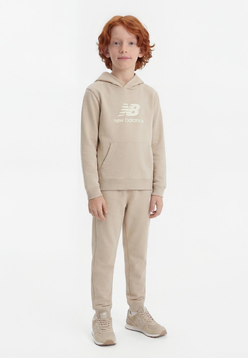 Παιδί με κοκκινομάλλες που φοράει μπεζ φούτερ New Balance, ταιριαστό παντελόνι φόρμας και αθλητικά παπούτσια, όρθιο μπροστά σε λευκό φόντο.