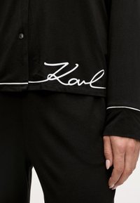 Črna srajca na gumb z belim obrobjem, na robu pa je napis "Karl" v kurzivnem fontu. Narejena iz gladkega materiala, ponuja sproščen kroj.