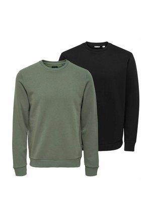 Junger Mann mit lockigem Haar, der einen olivgrünen Pullover, schwarze Hose und braune Schuhe trägt, steht und schaut zur Seite auf einem weißen Hintergrund.