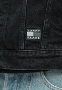 Μαύρο τζιν μπουφάν με υφασμένη ετικέτα που φέρει το κείμενο "TOMMY JEANS". Φθαρμένο τελείωμα και υφή στο ύφασμα. Μερικά απ ό τα ανοιχτόχρωμα μπλε τζιν είναι ορατά.