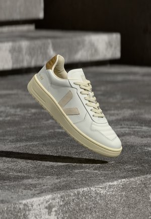 Baskets blanches et beiges avec "V-88" sur le talon, logo "V" beige et lacets, flottant au-dessus d’un fond de marches en béton texturé.