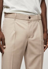 Beżowe spodnie wykonane z gładkiego materiału, z przednimi plisami, talią zapinaną na pojedynczy guzik oraz bocznymi kieszeniami.