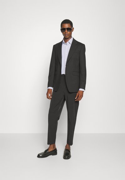 Férfi fekete öltönyben, világoskék ingben, fekete loaferekben és sötét napszemüvegben áll egy sima fehér háttér előtt.