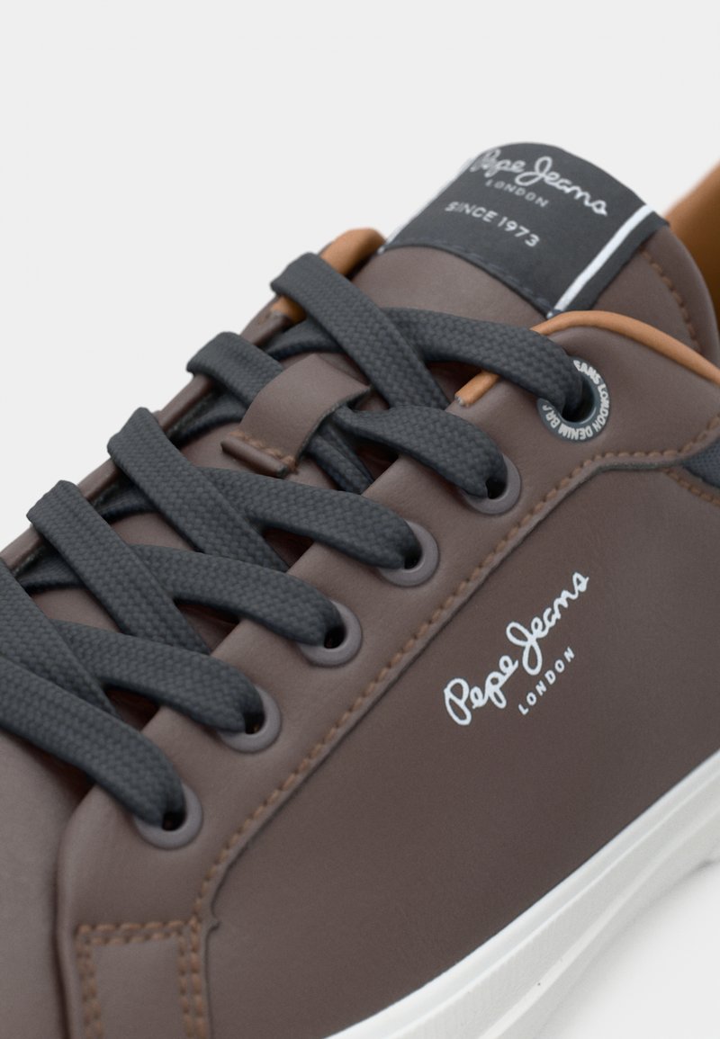 Zapatillas de piel sintética marrón con cordones grises, suela blanca, logo de la marca en el lateral y detalles en contraste en color marrón cerca de los ojetes.