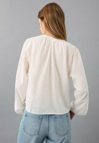 Blusa blanca de manga larga con acabado texturizado. Presenta un escote fruncido y un ajuste holgado, combinada con jeans de mezclilla azul claro.