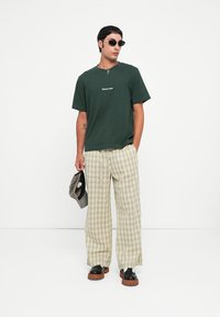 Tricou din bumbac verde închis cu text alb brodat, asortat cu pantaloni largi în carouri verzi deschise și pantofi cu platformă groasă negri.