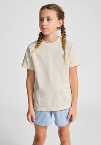 Lys beige bomulds t-shirt med korte ærmer og rund halsudskæring. Har et lille blåt pilgrafik på brystet. Kombineret med lyseblå shorts.