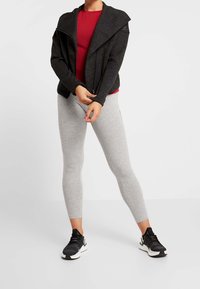 Šedé legíny, červený přiléhavý top a černá asymetrická bunda se zipem vyrobená z měkkého materiálu. Celý outfit doplňují černé tenisky.